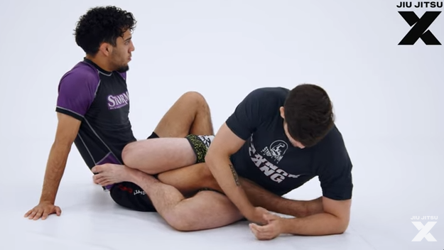 Two grapplers sitting in front of one another with their legs entangled and one players foot trapped in the other player&rsquo;s armpit.