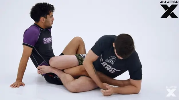 Two grapplers sitting in front of one another with their legs entangled and one players foot trapped in the other player's armpit.